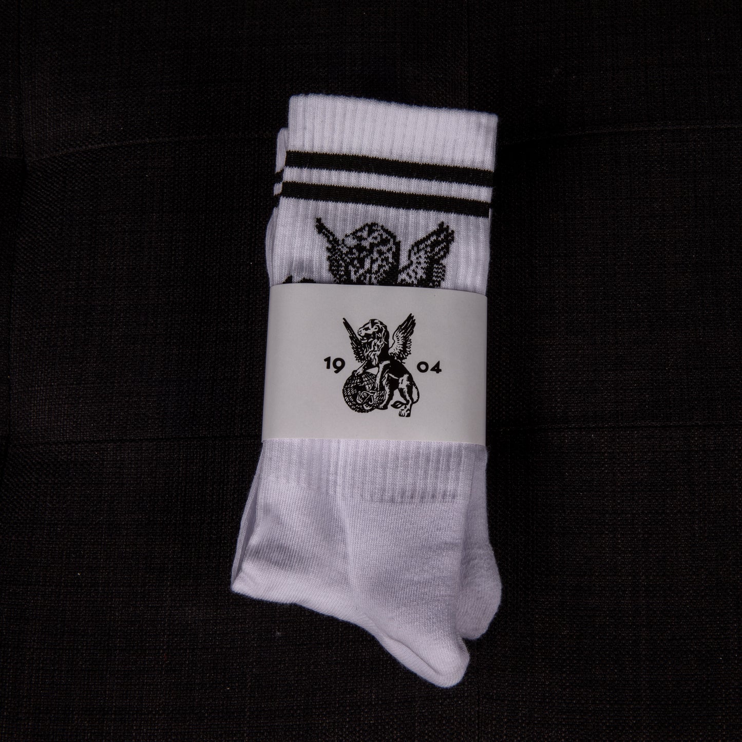 Weiße Socken mit schwarzen Streifen und aufgesticktem geflügeltem Löwen, in Verpackung von oben fotografiert – mit 1904-Emblem.