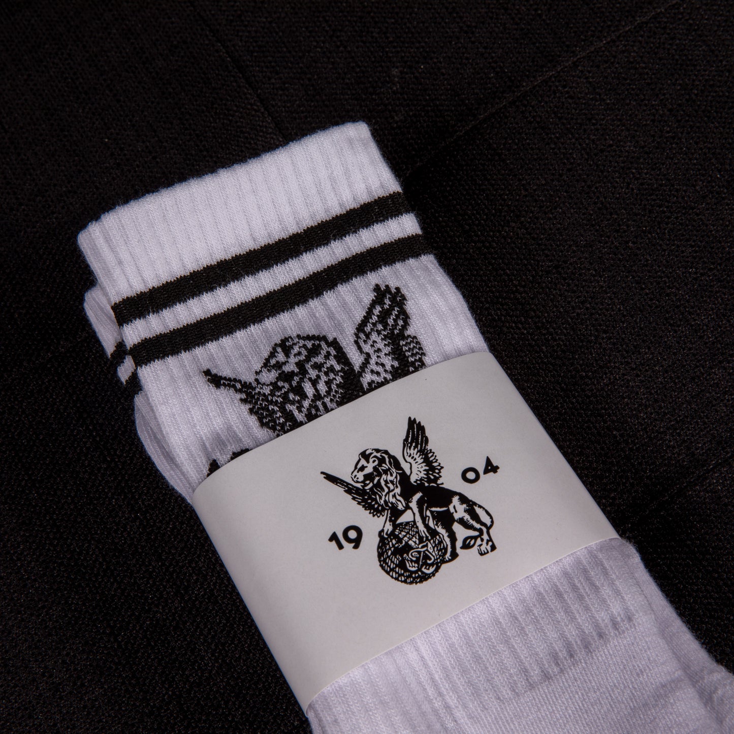 Weiße Sportsocken mit schwarzen Streifen und Löwenmotiv, in Verpackung mit geflügeltem Löwenlogo und der Jahreszahl 1904 – Vorderansicht.