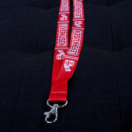 Rotes Lanyard mit weißen TSV-Bayer-04-Leverkusen-Logos und Schriftzug ‚Leverkusen – mit dem Kreuz auf der Brust‘, mit silbernem Karabinerhaken, flach auf dunklem Untergrund fotografiert.