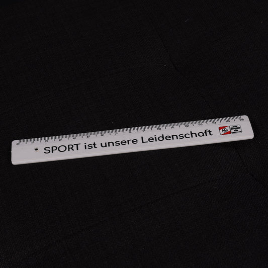 Weißes Kunststofflineal mit schwarzer Aufschrift ‚SPORT ist unsere Leidenschaft‘ und TSV-Bayer-04-Leverkusen-Logo, auf dunklem Untergrund liegend.