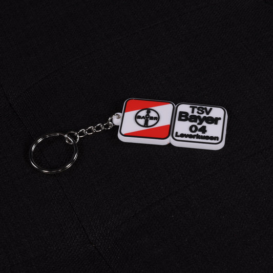 Gummierter Schlüsselanhänger mit Kette und Ring, im Design des TSV Bayer 04 Leverkusen Logos in Rot, Weiß und Schwarz, liegend auf schwarzem Stoff.