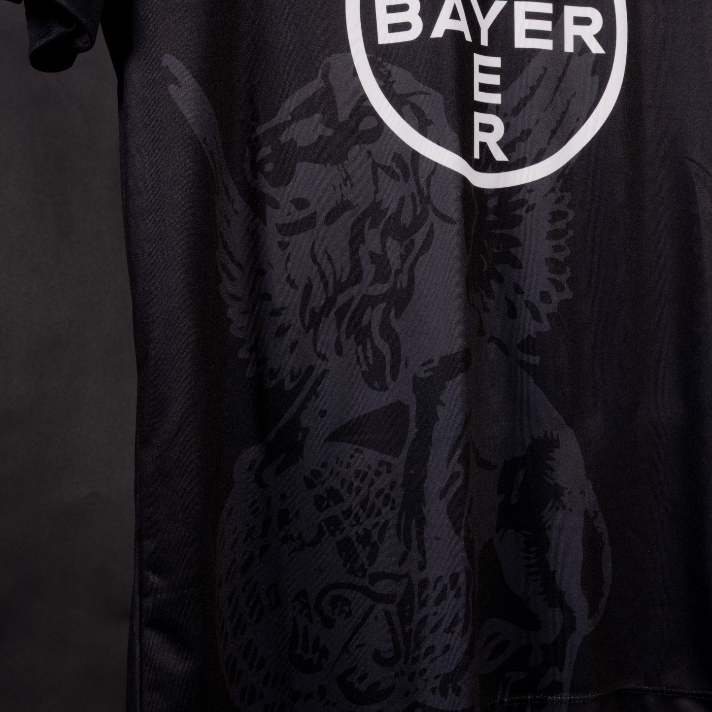 Detailansicht des unteren Bereichs des schwarzen Sondertrikots von TSV Bayer 04 Leverkusen mit dem weißen Bayer-Kreissymbol und großflächigem, dunkel eingearbeitetem Löwenmotiv.