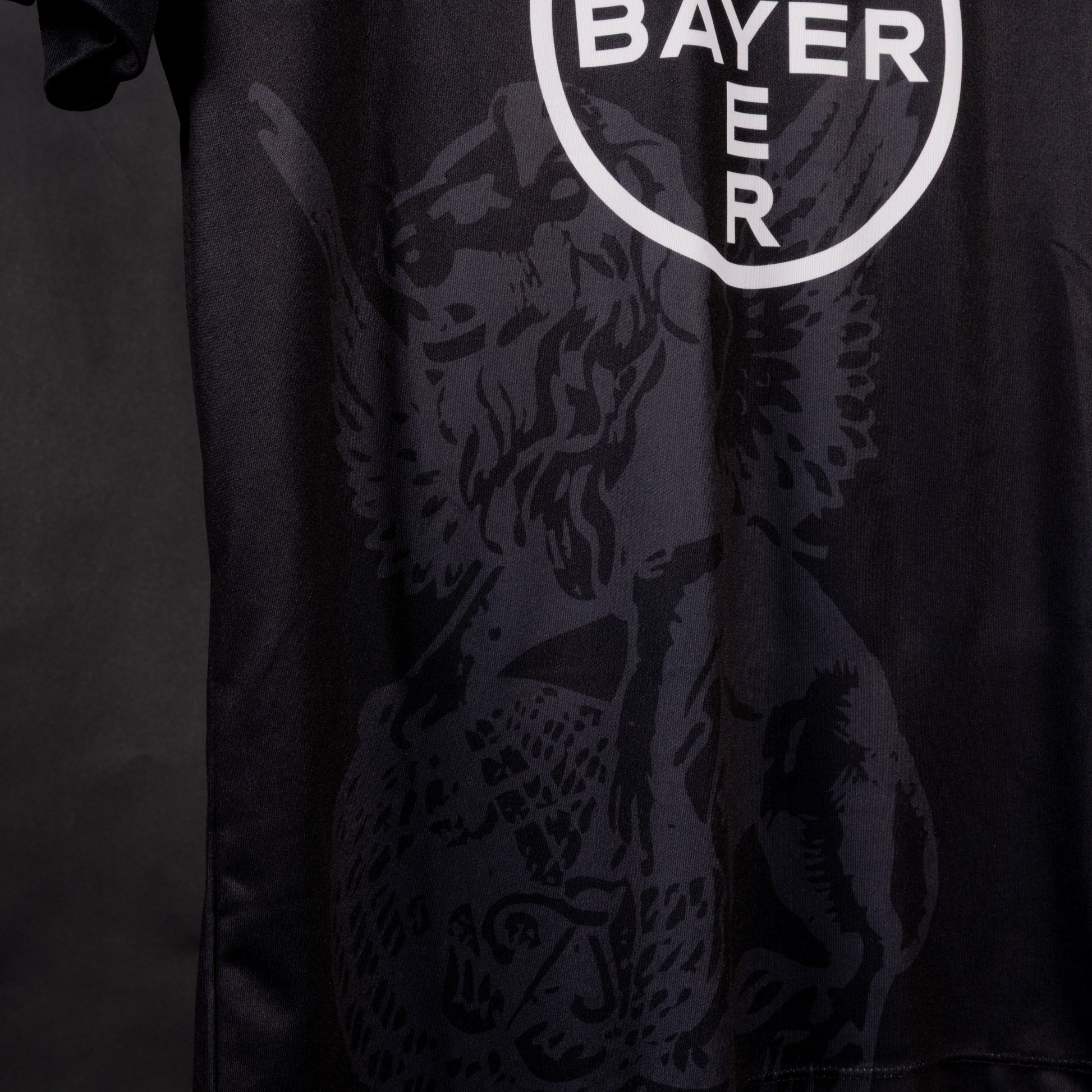 Detailansicht des unteren Bereichs des schwarzen Sondertrikots von TSV Bayer 04 Leverkusen mit dem weißen Bayer-Kreissymbol und großflächigem, dunkel eingearbeitetem Löwenmotiv.