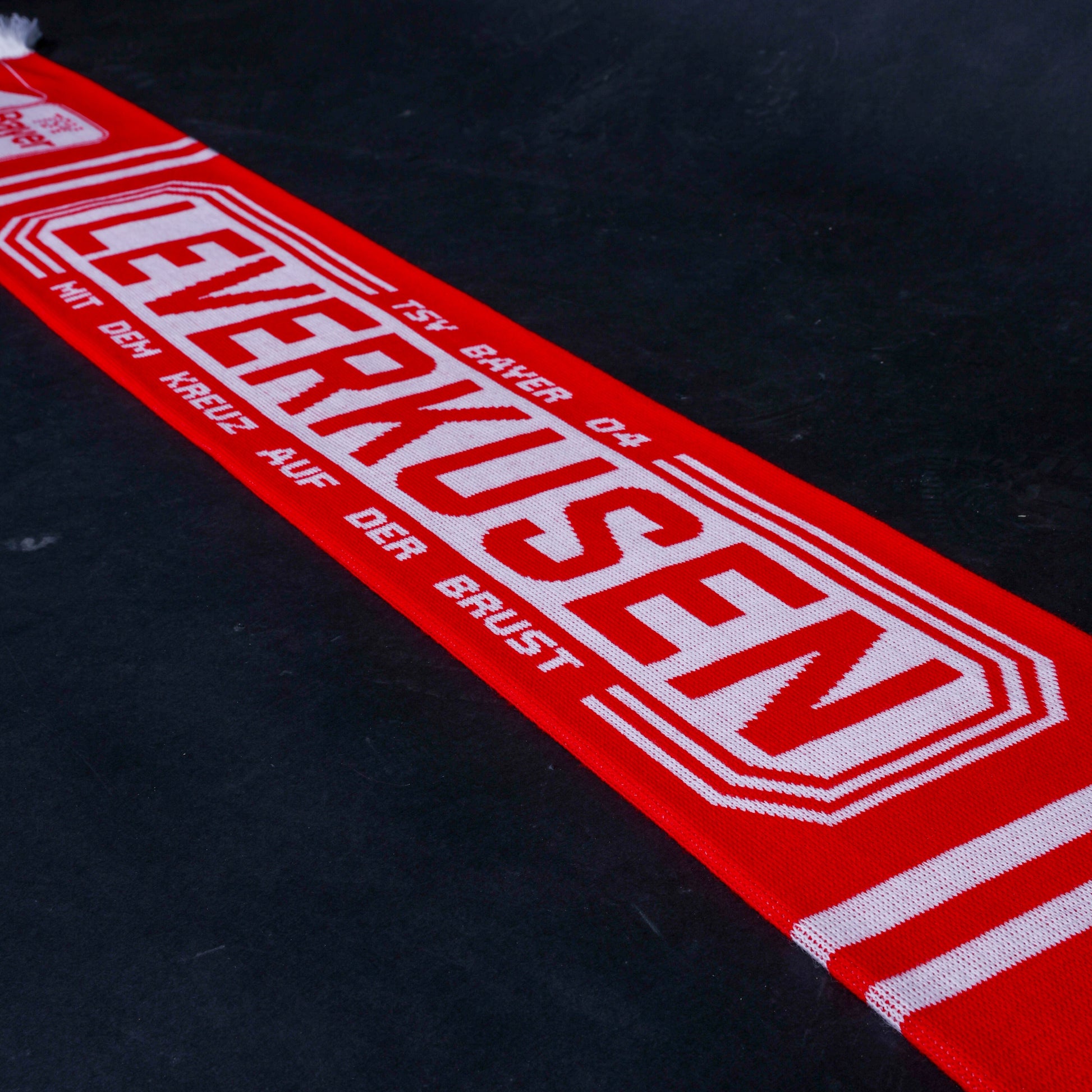 Roter TSV-Bayer-04-Leverkusen-Fanschal in Draufsicht auf dunklem Untergrund, mit großem weißen Schriftzug ‚LEVERKUSEN – MIT DEM KREUZ AUF DER BRUST‘ und weißer Doppelumrandung.