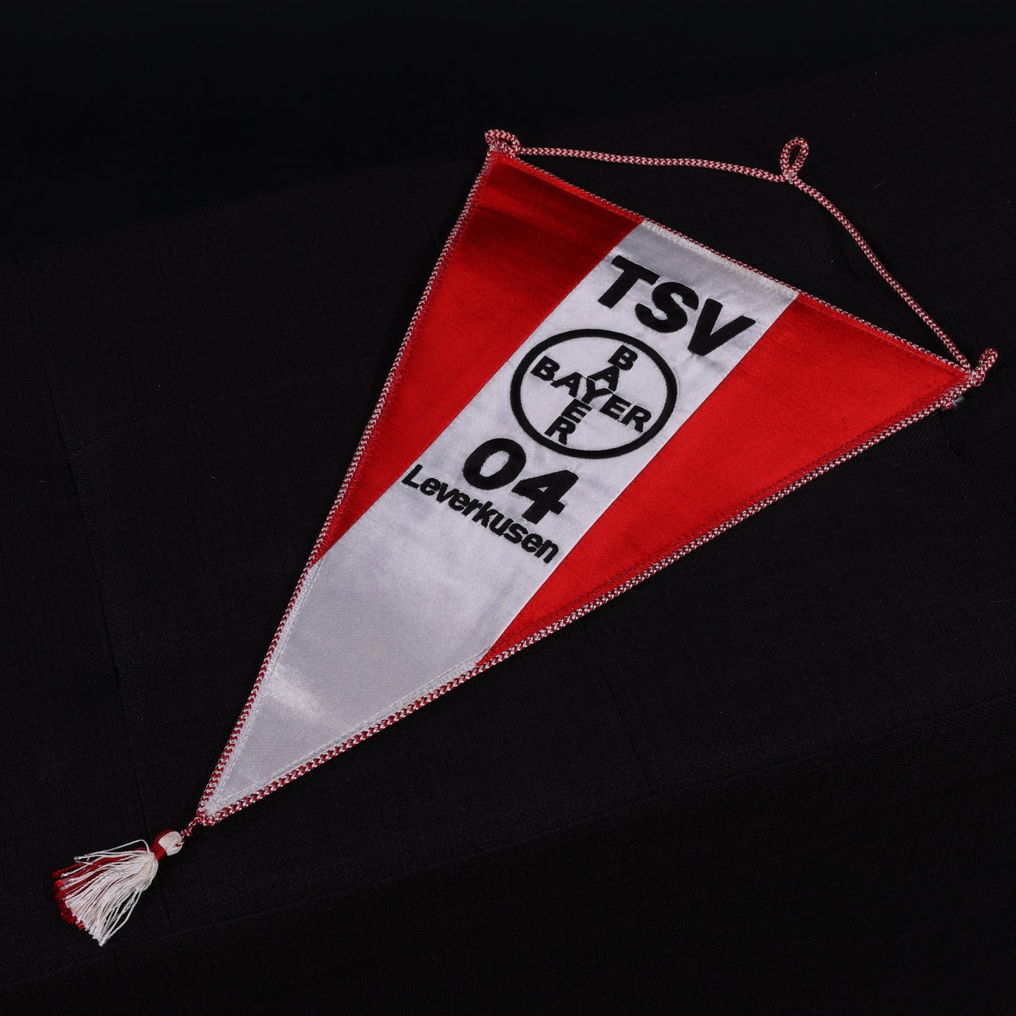 Dreieckiger Stoffwimpel in Rot-Weiß mit Aufdruck ‚TSV Bayer 04 Leverkusen‘ und rundem Vereinslogo, mit Kordelaufhängung und Fransen, liegend auf schwarzem Untergrund.