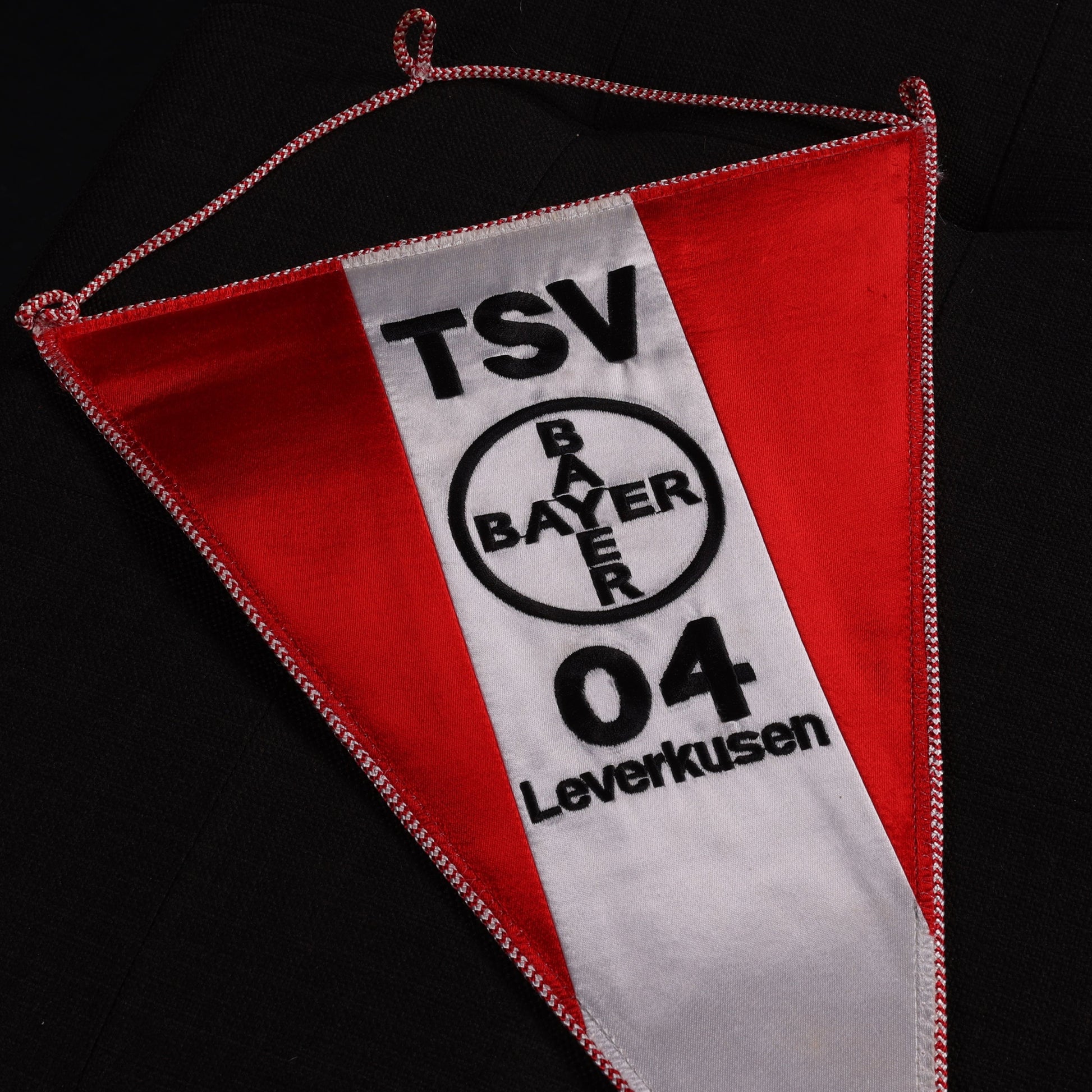 Nahaufnahme des oberen Bereichs eines TSV-Bayer-04-Leverkusen-Wimpels mit rotem und weißem Stoff, schwarzem Vereinslogo und Schriftzug.