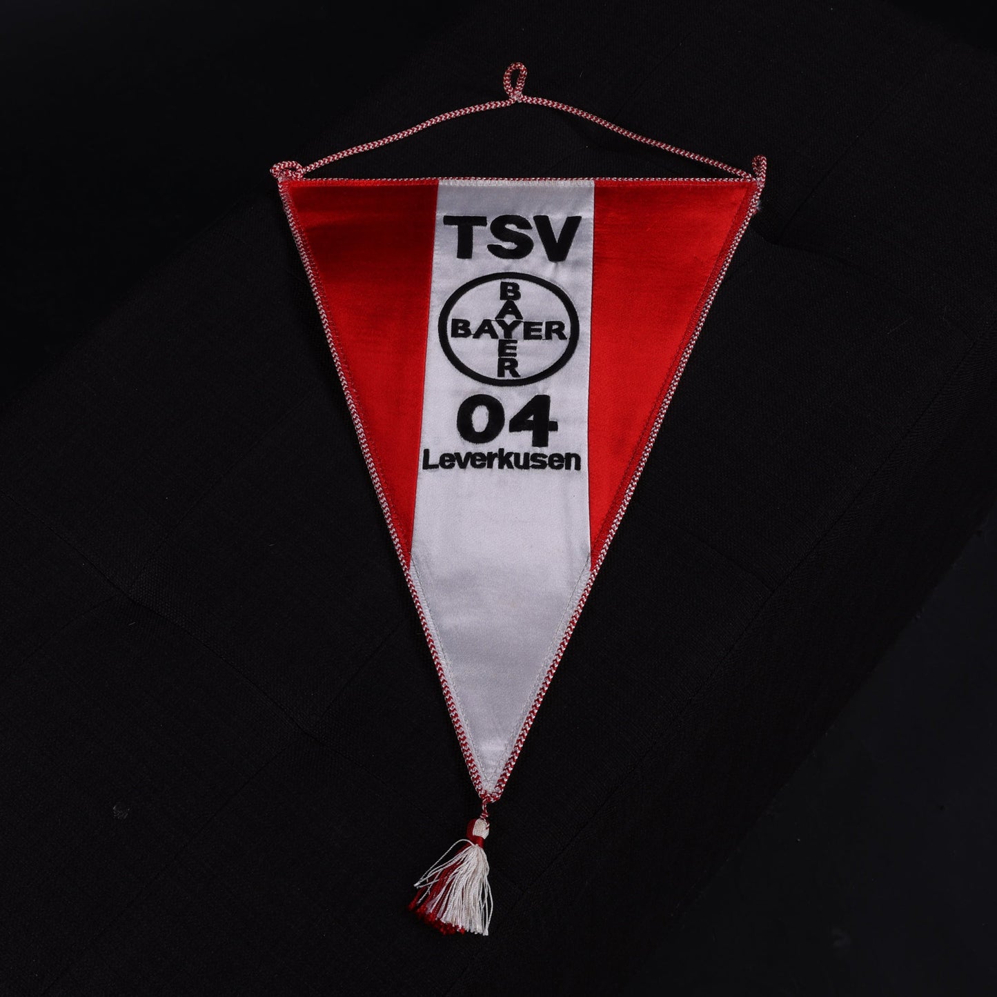 Zentral fotografierter Wimpel des TSV Bayer 04 Leverkusen in Rot-Weiß mit Vereinslogo und Schriftzug, auf schwarzem Untergrund ausgebreitet.