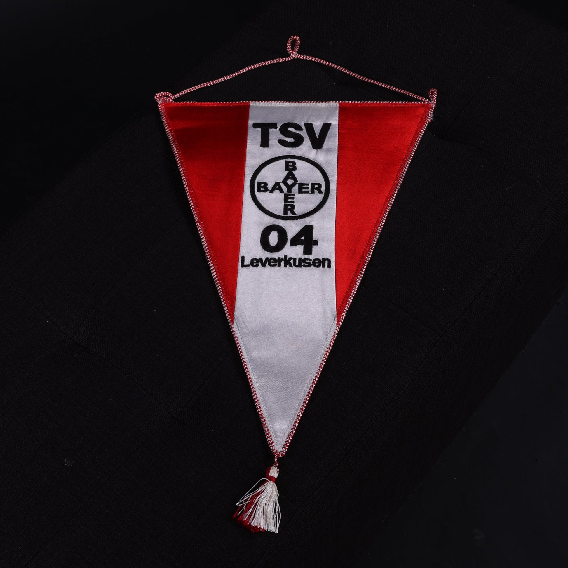 Zentral fotografierter Wimpel des TSV Bayer 04 Leverkusen in Rot-Weiß mit Vereinslogo und Schriftzug, auf schwarzem Untergrund ausgebreitet.
