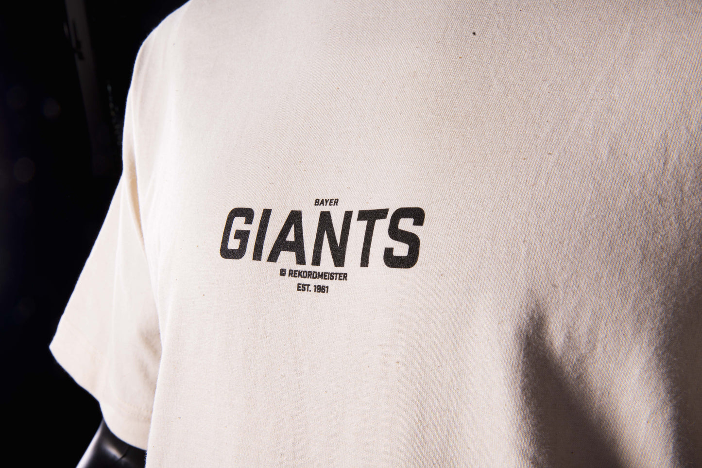 Nahaufnahme des Brustprints auf dem beigefarbenen Bayer-Giants-T-Shirt mit schwarzer Aufschrift ‚Bayer Giants – Rekordmeister – Est. 1961'.