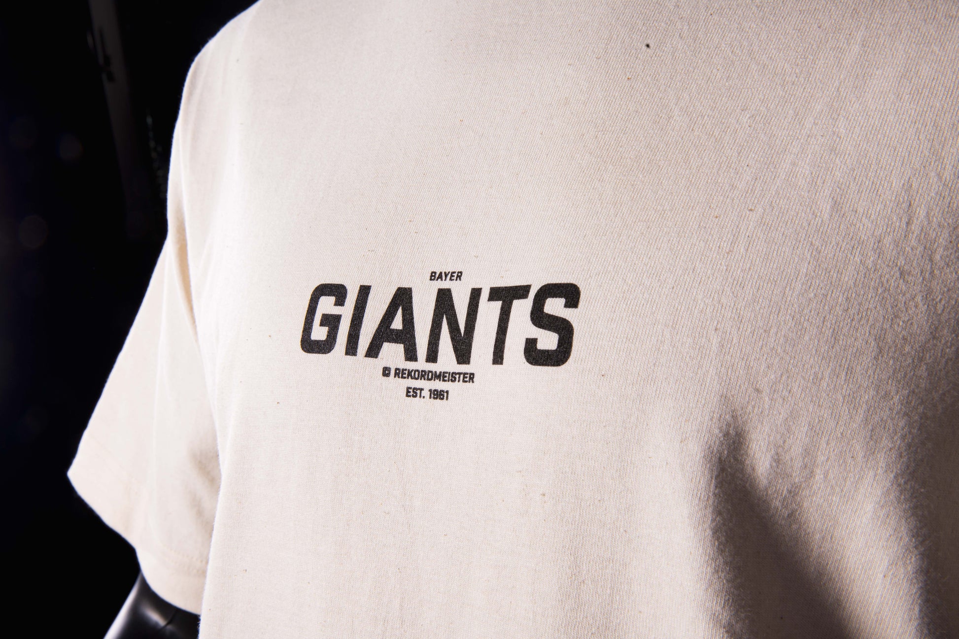 Nahaufnahme des Brustprints auf dem beigefarbenen Bayer-Giants-T-Shirt mit schwarzer Aufschrift ‚Bayer Giants – Rekordmeister – Est. 1961'.
