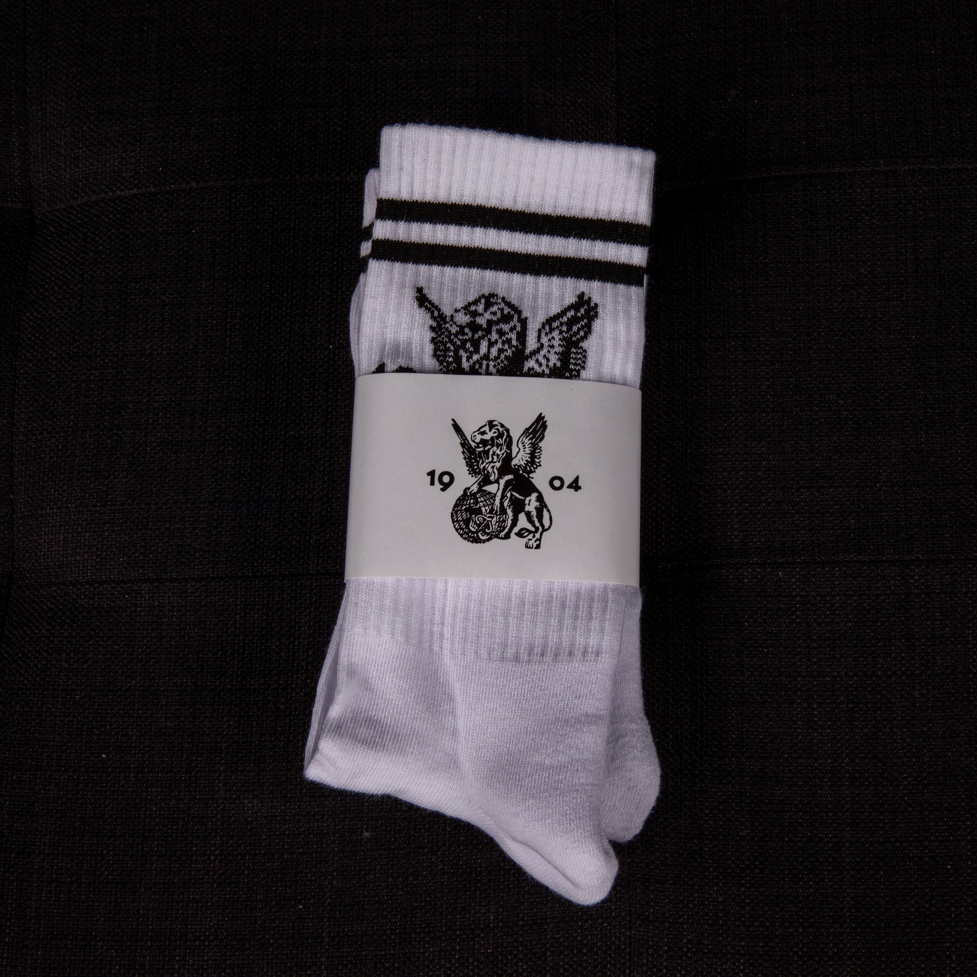 Weiße Socken mit schwarzen Streifen und aufgesticktem geflügeltem Löwen, in Verpackung von oben fotografiert – mit 1904-Emblem.